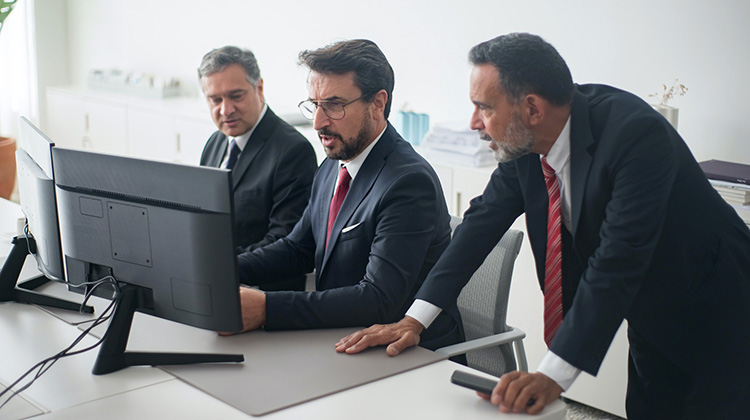 Tres hombres de negocios están sentados frente a un escritorio. Los tres argumentan sobre el contenido de finanzas que se muestra en un monitor. El escritorio es de una sala de juntas.